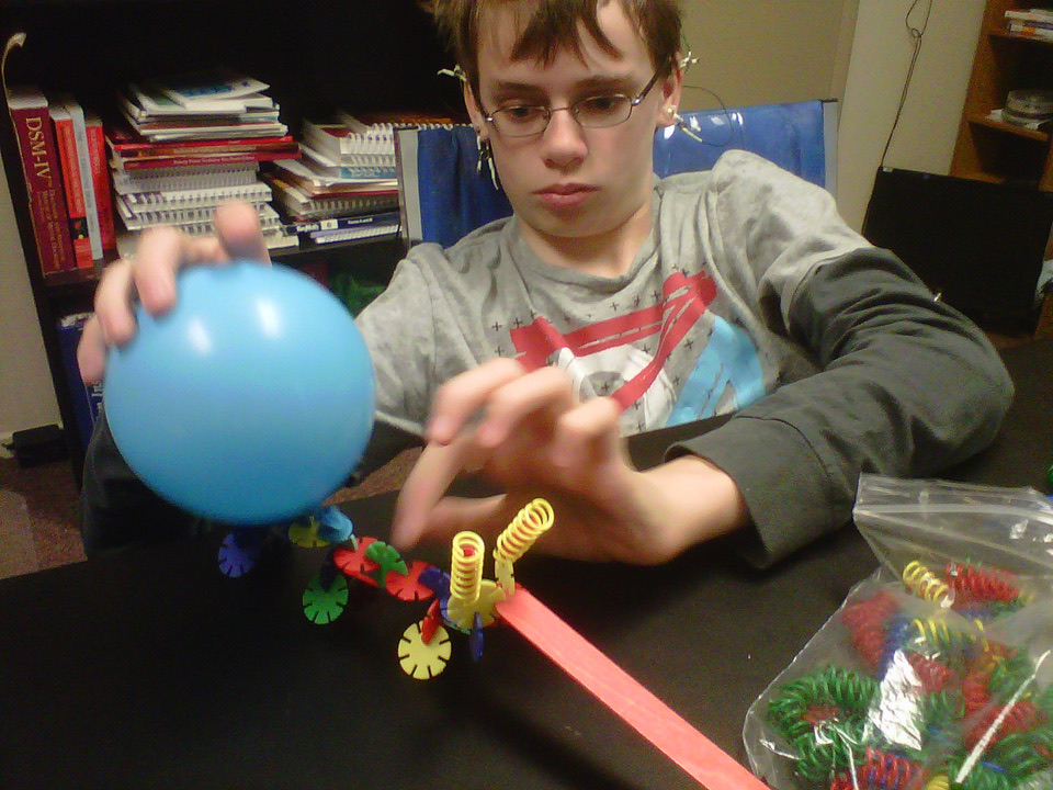 A teenage boy at a table working through a balloon and coil coordination exercise during a session.